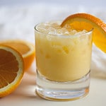 orange adrenal cocktail in a glass, garnished with an orange slice, and a sliced orange in the background