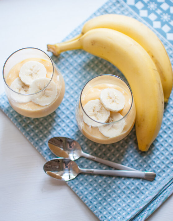 two glasses of 3-ingredient banana pudding next to two bananas and two spoons all on a blue placemat