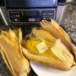 air fryer corn on the cob in husk on a white plate next to air fryer