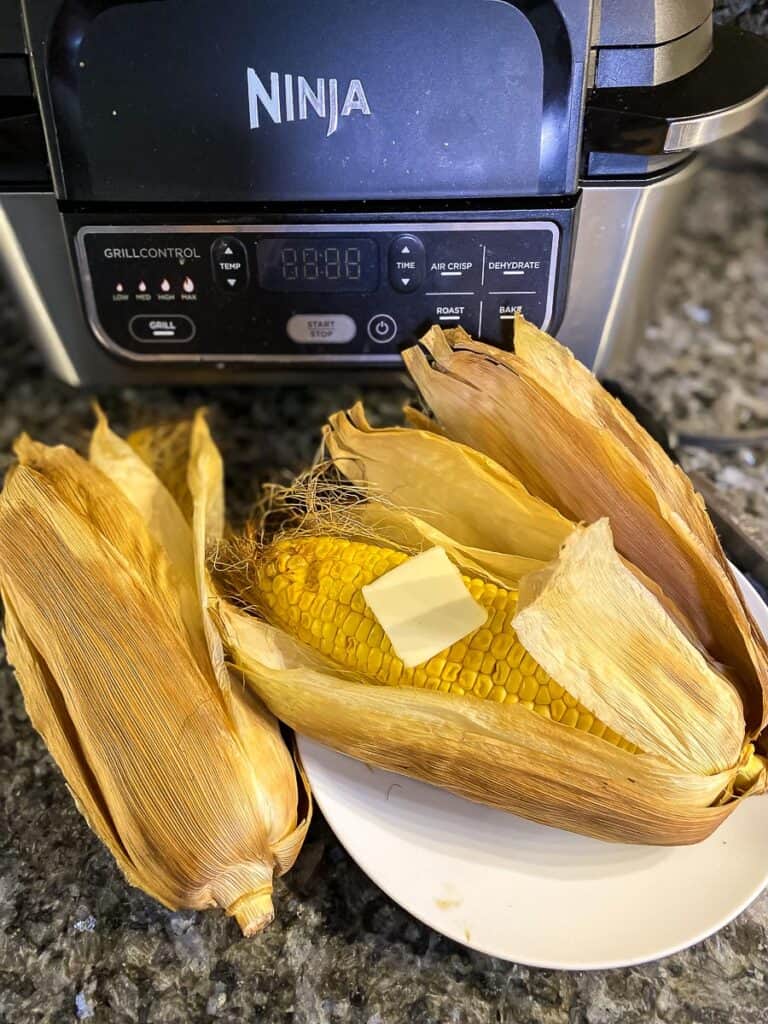 air fryer corn on the cob in husk on a white plate next to air fryer