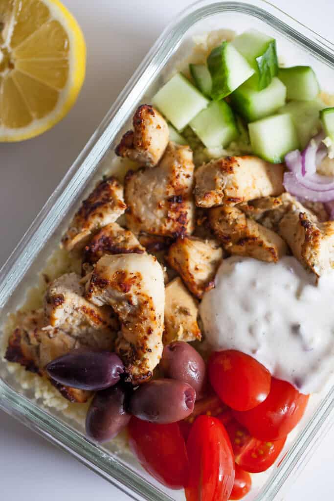 greek chicken bites in a meal prep container with sauce, tomatoes, cucumbers and lemon